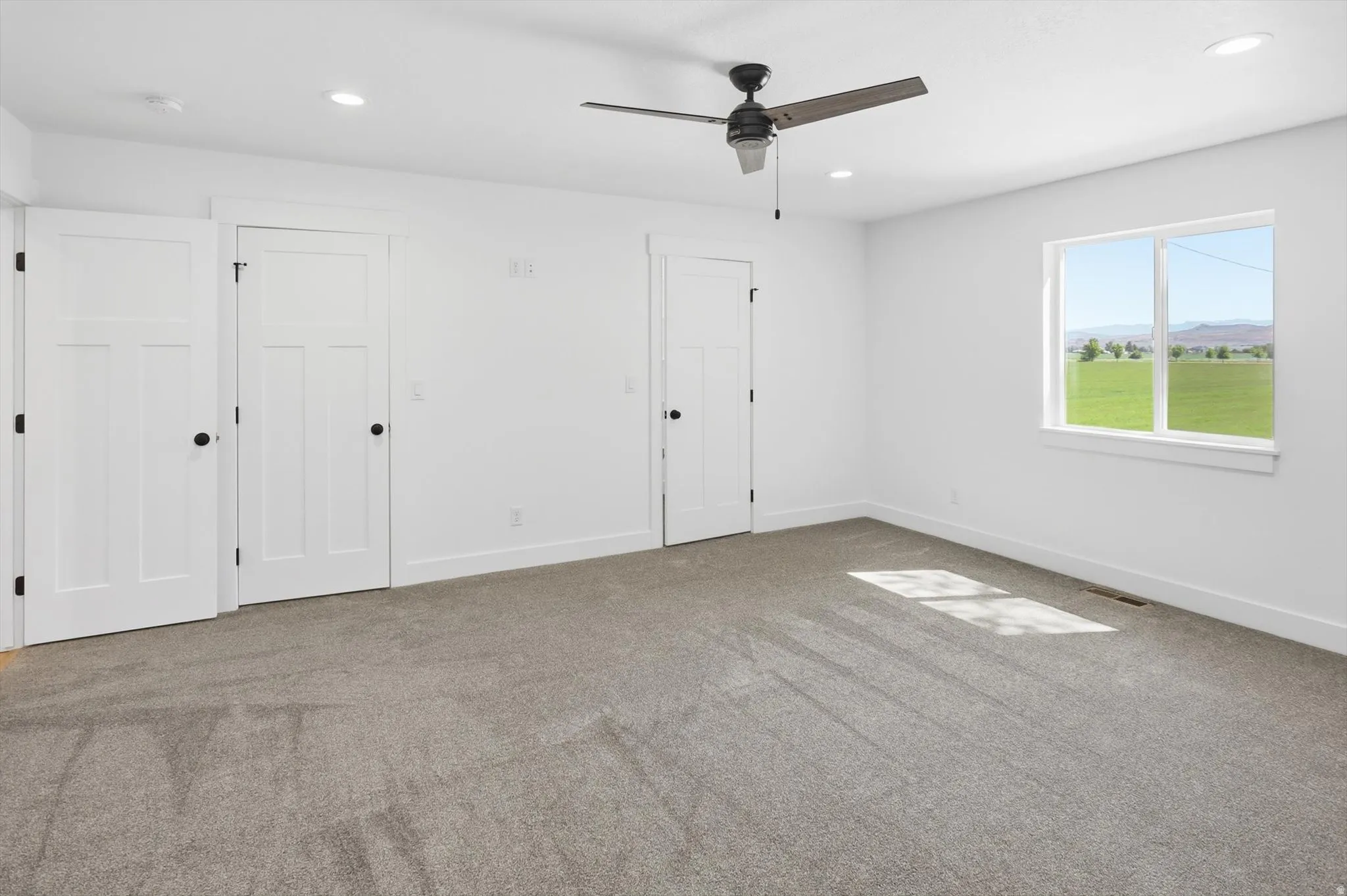 Spacious room featuring neutral carpeting, white walls, a ceiling fan, and a large window with a scenic landscape