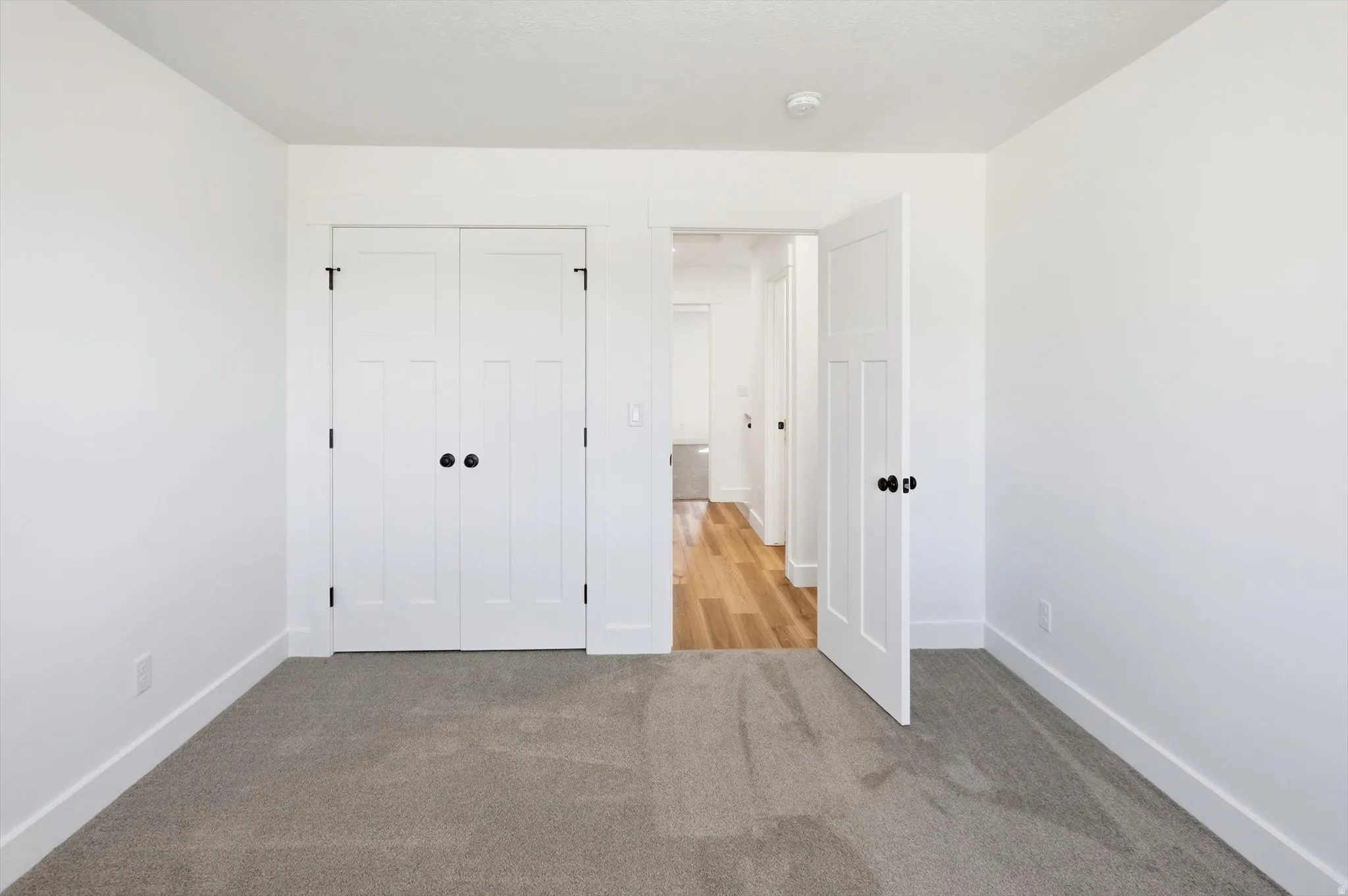 Spacious room featuring neutral carpeting, white walls, and a pair of white paneled closet doors with black hardware