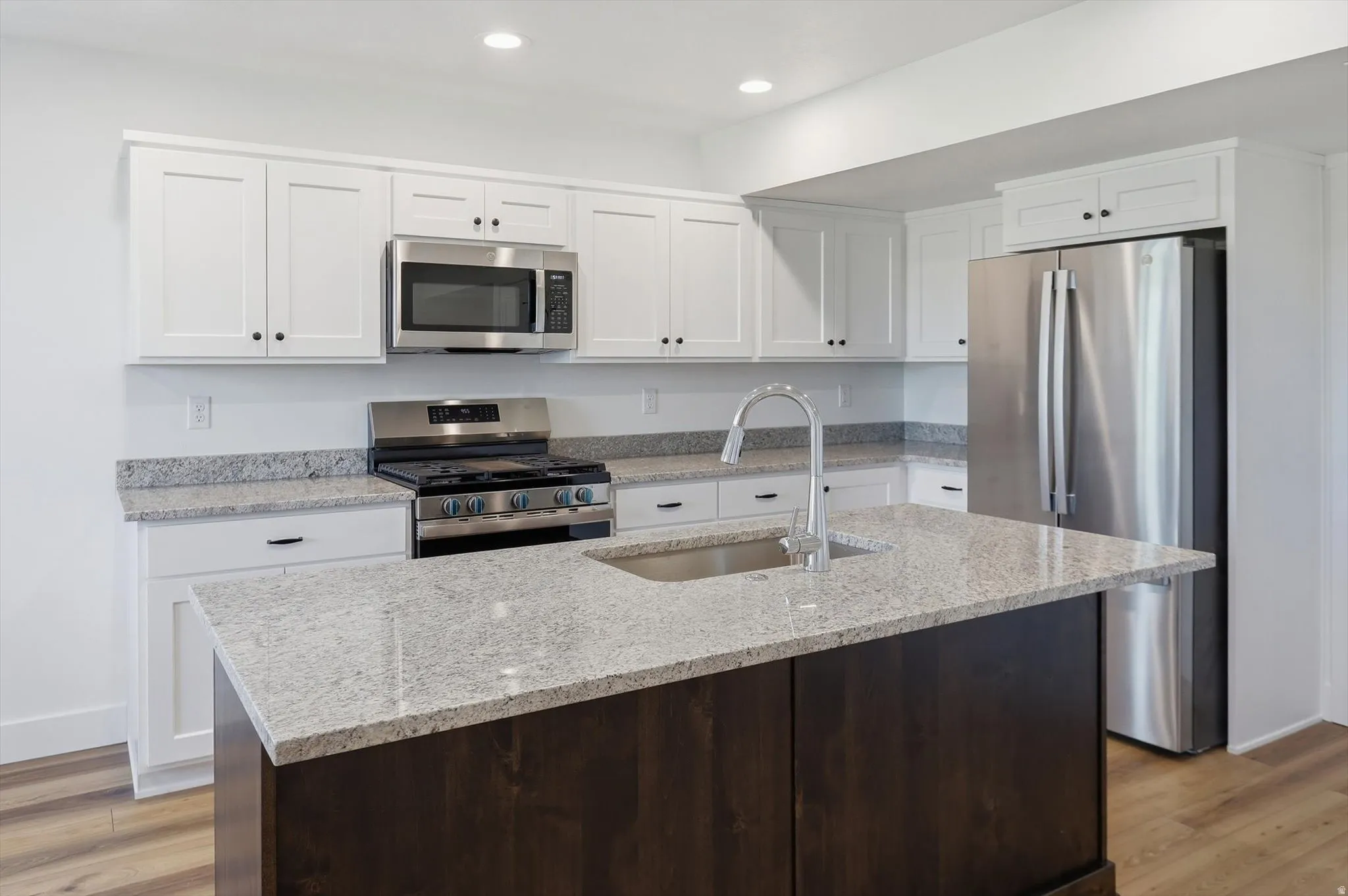 Kitchen featuring a central island with an undermount sink and gooseneck faucet, white shaker-style cabinetry with matte black hardware, stainless steel appliances, and wood-finish flooring