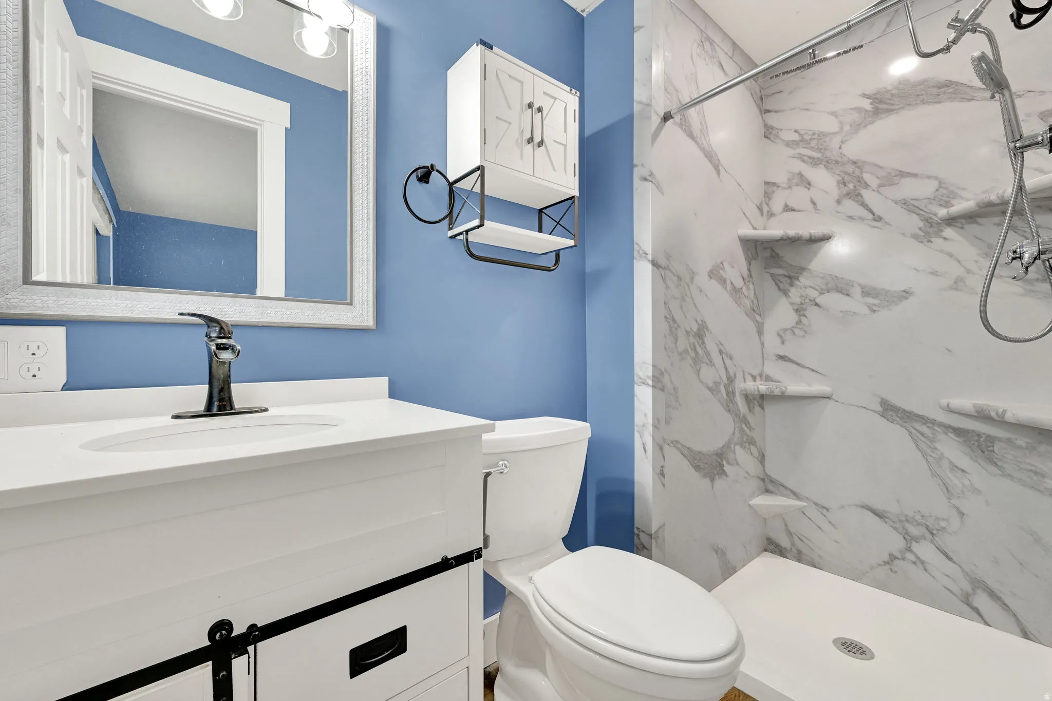 Bathroom featuring a walk-in shower with marble-look wall panels and built-in shelving, a white vanity with a single basin sink and black faucet, a framed mirror, and a wall-mounted storage cabinet
