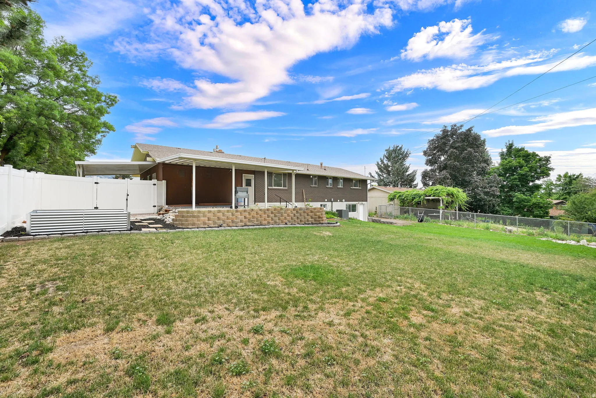 Expansive backyard featuring a manicured lawn, a white privacy fence, and a covered patio with a retaining wall
