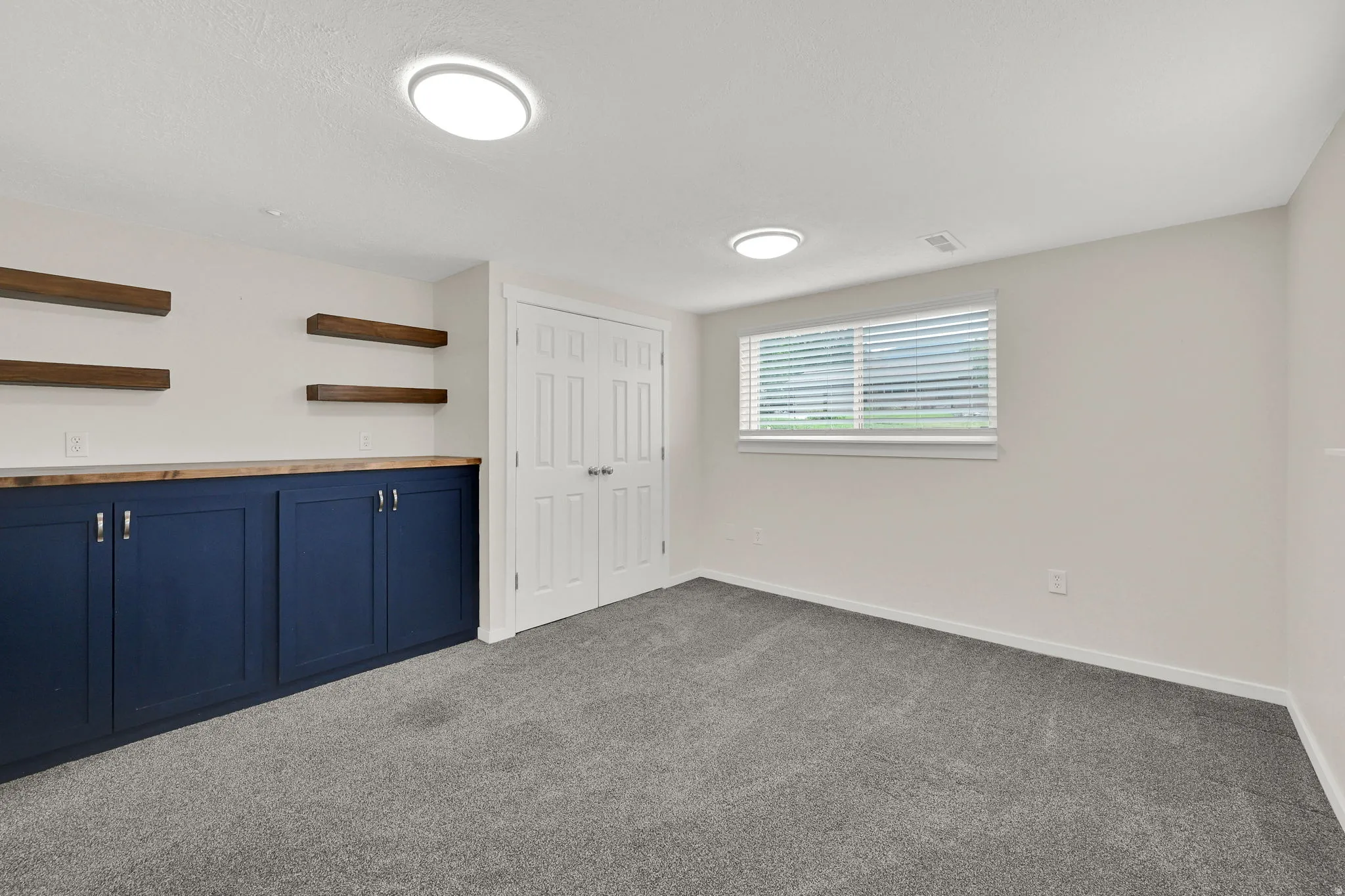 Finished room featuring built-in navy cabinetry with wood-finish countertop, floating wood-finish shelving, and neutral gray carpeting