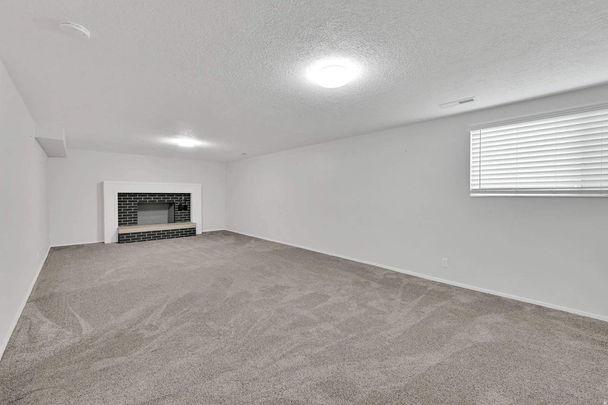 Spacious room with a brick-clad fireplace and hearth, neutral carpet, white walls, and a window with blinds