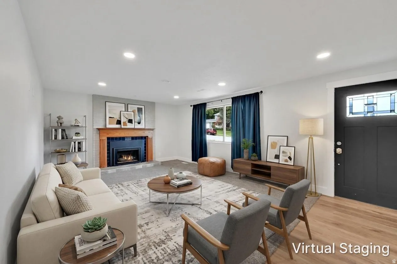 Living area featuring a fireplace with a wood mantle and tile surround, recessed lighting, wood-finish flooring, a contemporary dark entry door with a transom window, and windows with dark-colored curtains. Room was virtually staged.