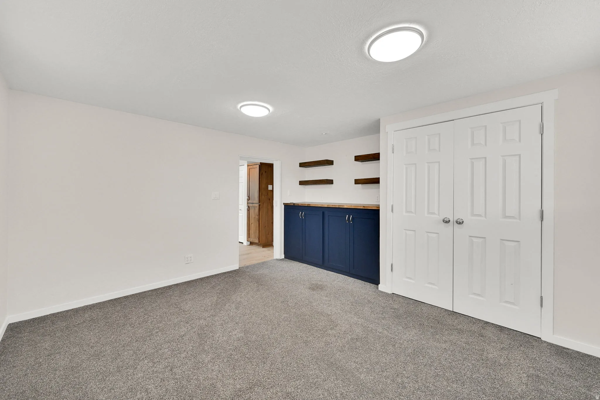 Room featuring plush carpeting, a built-in blue cabinet with a wood-finish countertop, three floating wood-finish shelves, and a double-door closet
