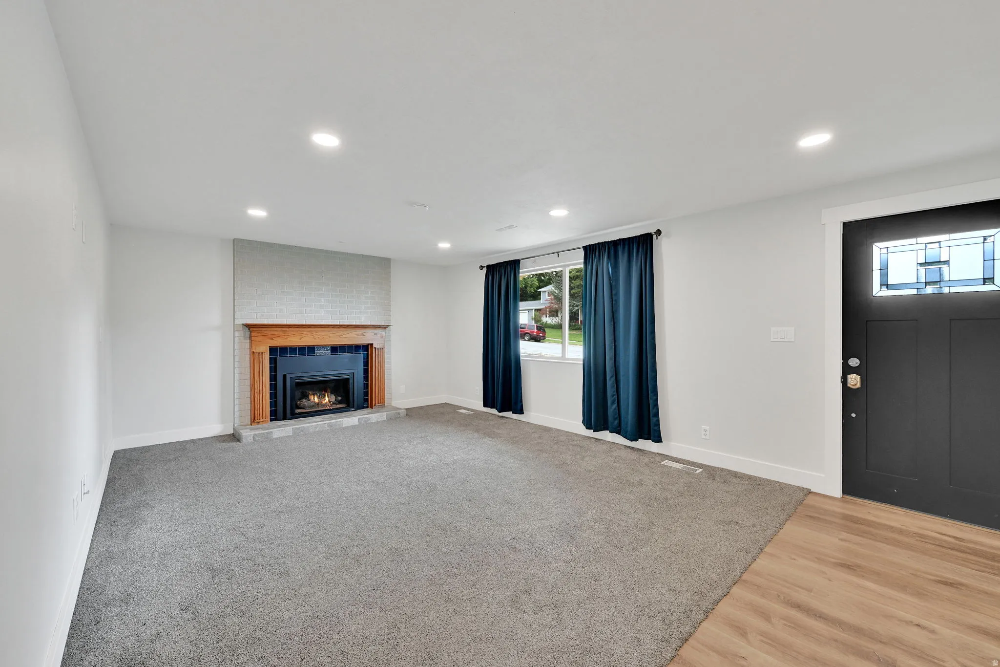 Neutral interior featuring a fireplace with a light brick surround and wood-finish mantel, recessed lighting, wood-finish flooring at the entry, and a dark paneled door with decorative glass