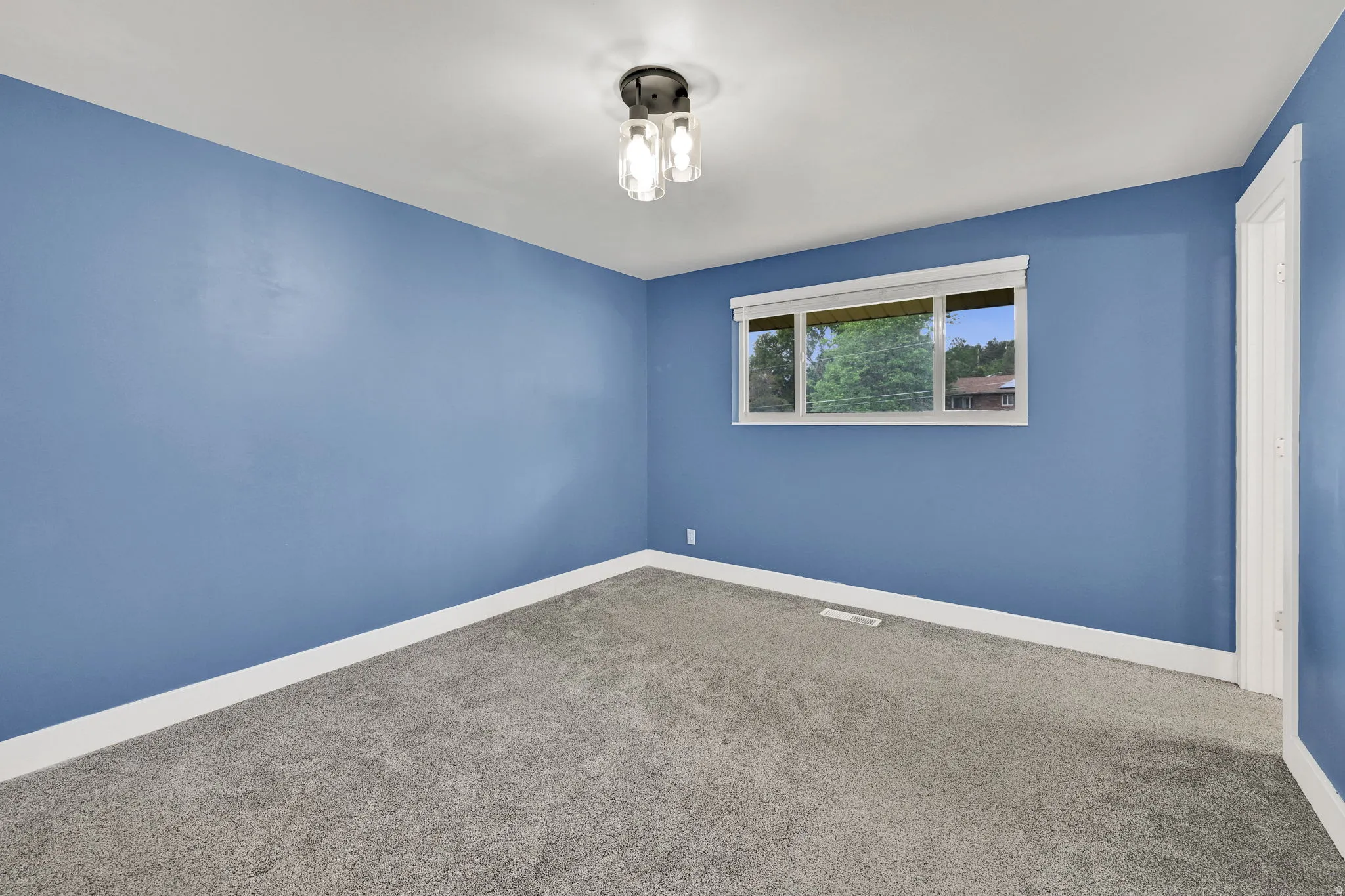 Spacious room with blue walls and light gray carpeting, featuring a multi-pane window with white trim, a modern three-light ceiling fixture, and white baseboards