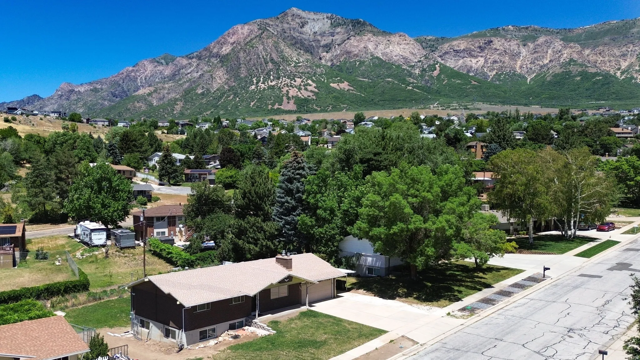Mountain backdrop showcasing a residential property with a brown-roofed structure, attached carport, concrete driveway, and established landscaping