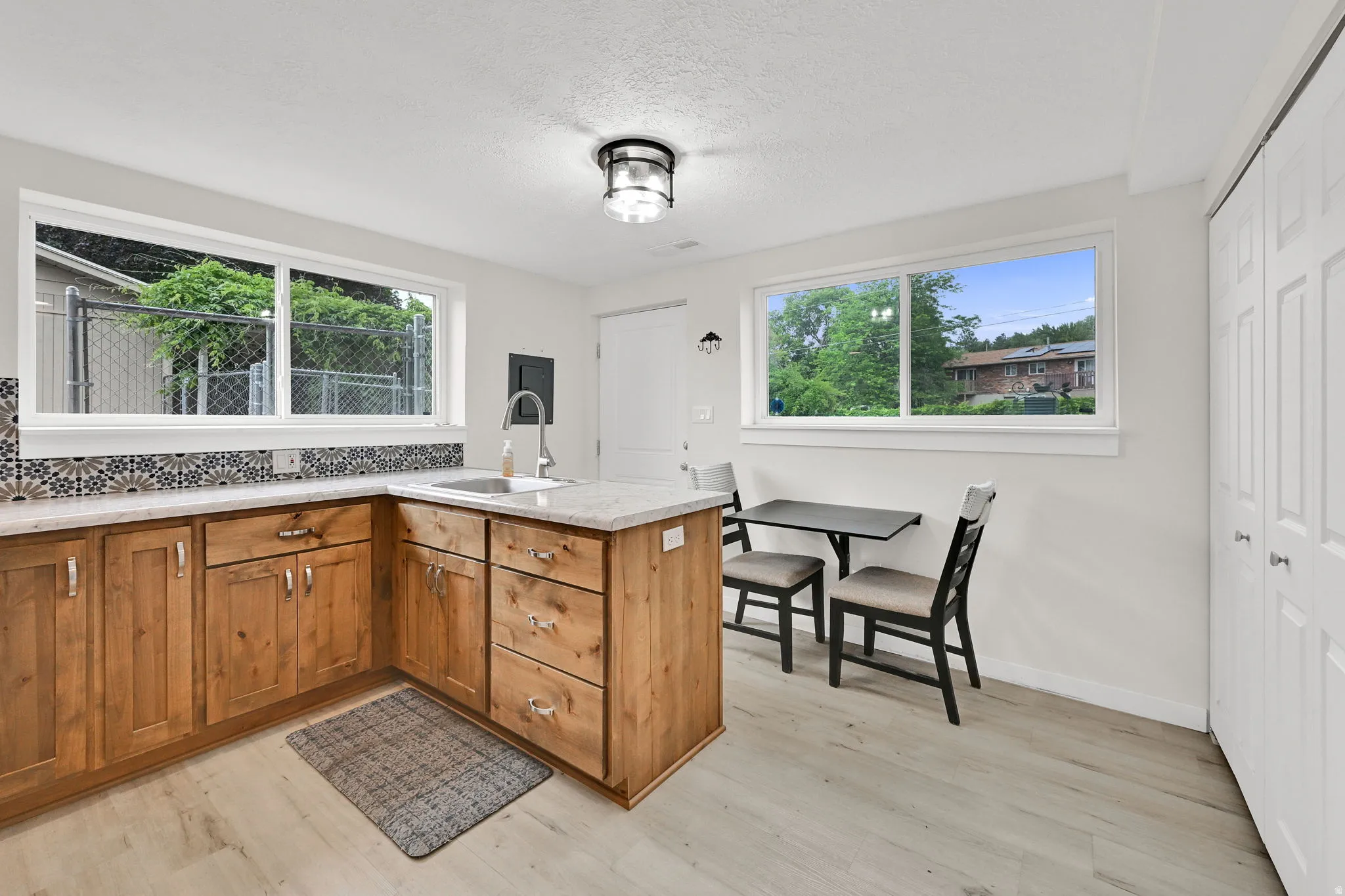 Light-filled kitchen featuring natural wood cabinetry, light-toned countertops, a patterned tile backsplash, wood-finish flooring, and a stainless steel sink with a high-arc faucet