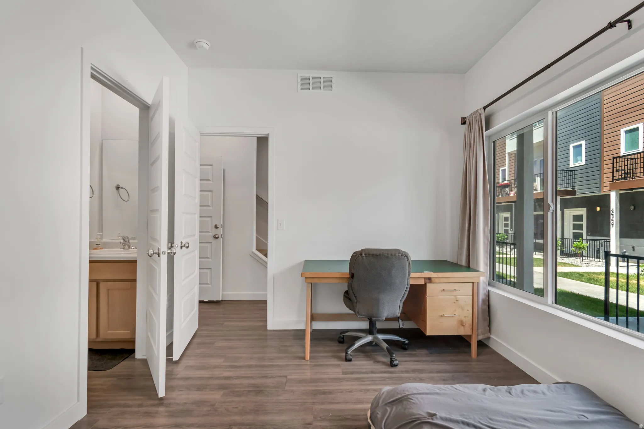 Spacious room with wood-finish flooring, a large window, white walls, and a visible doorway to an adjacent bathroom with a vanity