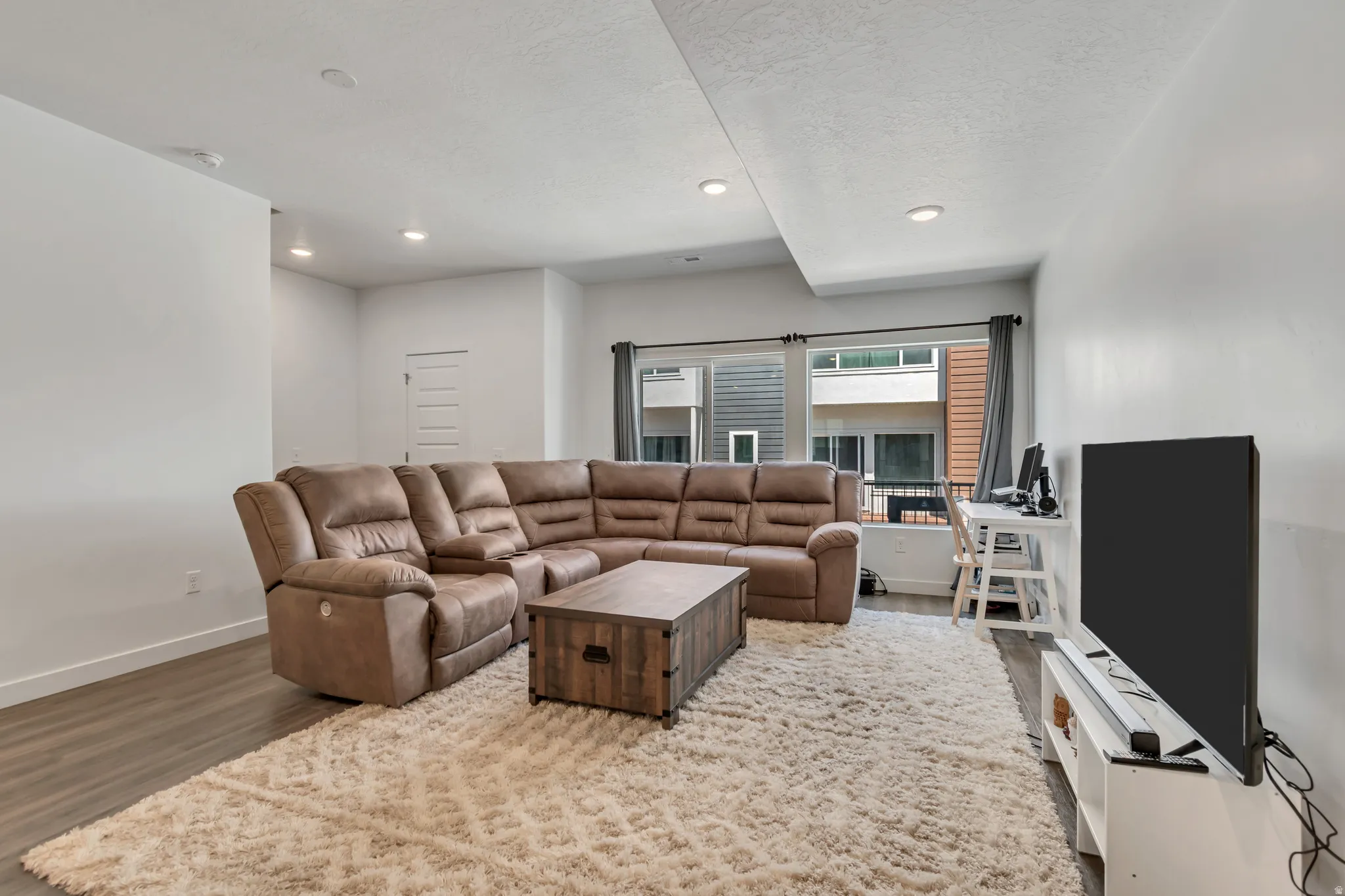 Spacious living area featuring light wood-finish flooring, recessed lighting, and a large window with privacy blinds