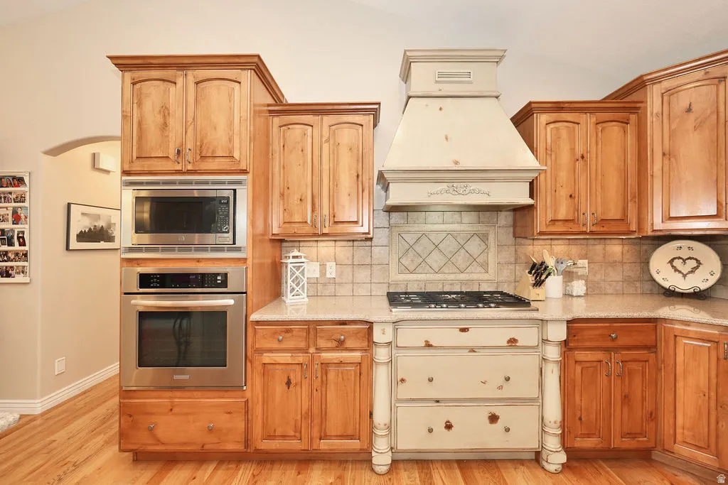 Kitchen featuring custom cabinetry with natural wood-finish and distressed cream finishes, a built-in stainless steel microwave and oven, a gas cooktop, and a decorative range hood