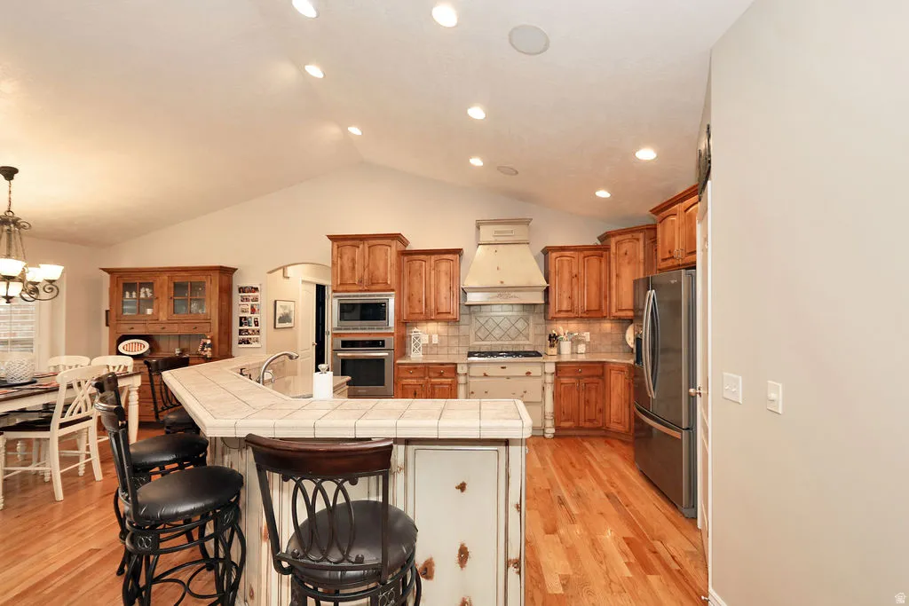 Kitchen featuring wood-finish flooring, vaulted ceiling with recessed lighting, tile-topped peninsula with seating, stainless steel appliances, and wood cabinetry