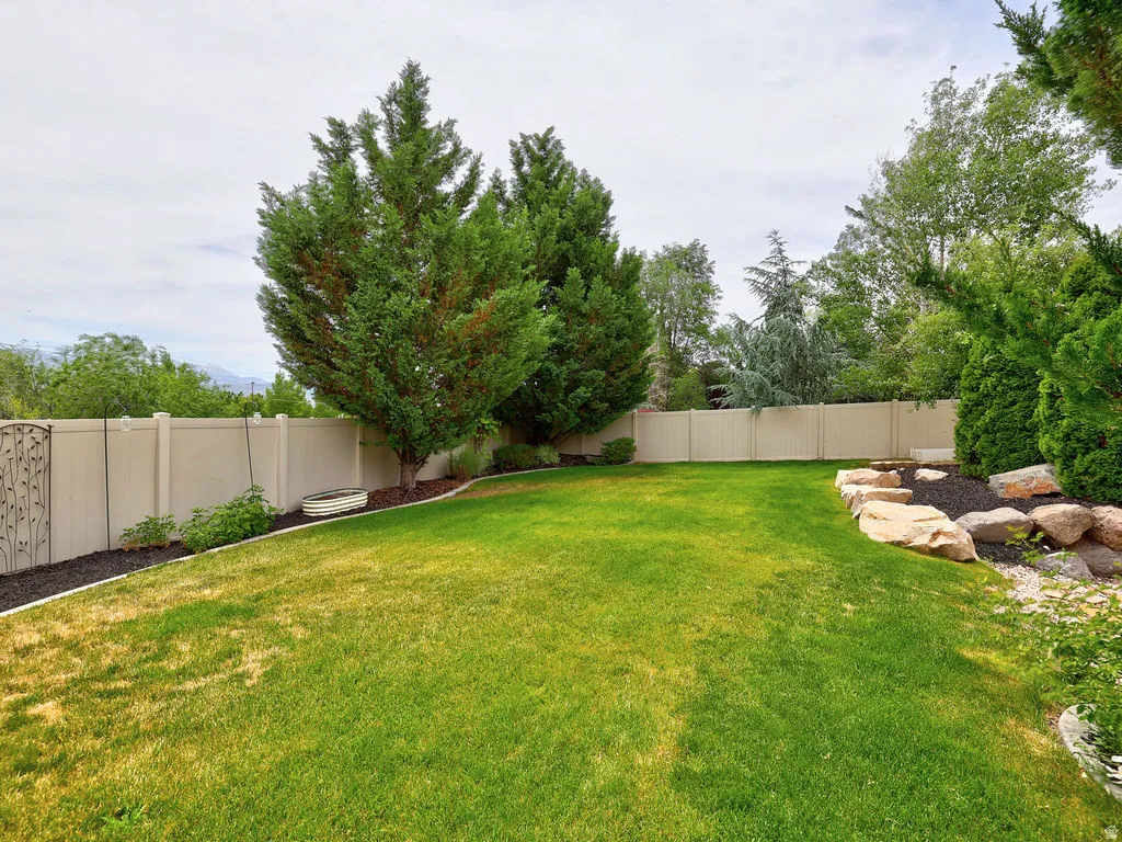 Expansive backyard featuring a lush lawn, mature trees, a durable privacy fence, a decorative metal gate, and a natural rock garden accent