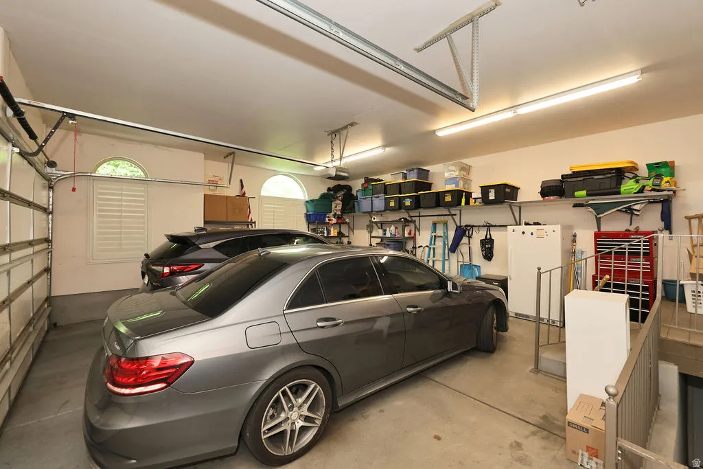 Spacious two-car garage featuring a concrete floor, arched windows with plantation shutters, overhead fluorescent lighting, and built-in wall shelving