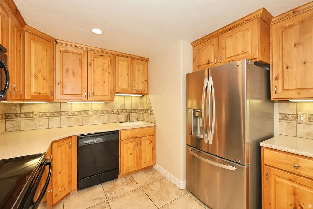 Kitchen featuring natural wood cabinetry, stainless steel refrigerator, black dishwasher, beige tile flooring, and light-toned countertops
