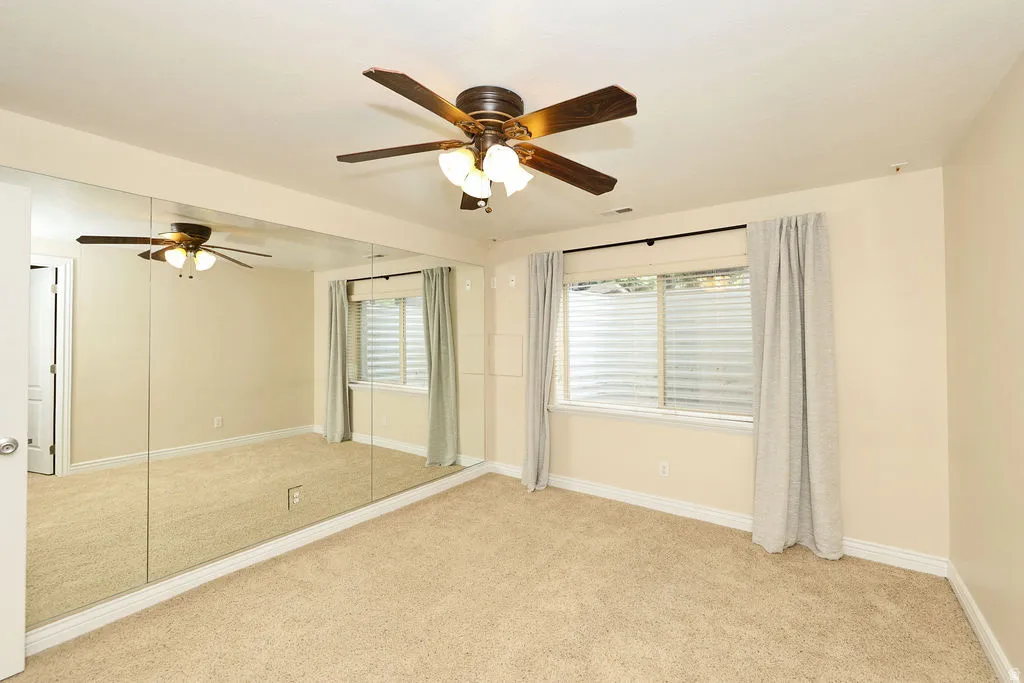 Carpeted room featuring a full wall of mirrors, a ceiling fan with integrated lighting, a window with horizontal blinds, and light-colored walls