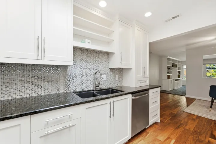 White cabinetry with silver-tone hardware, dark countertop, mosaic tile backsplash, double basin sink with gooseneck faucet, and stainless steel dishwasher