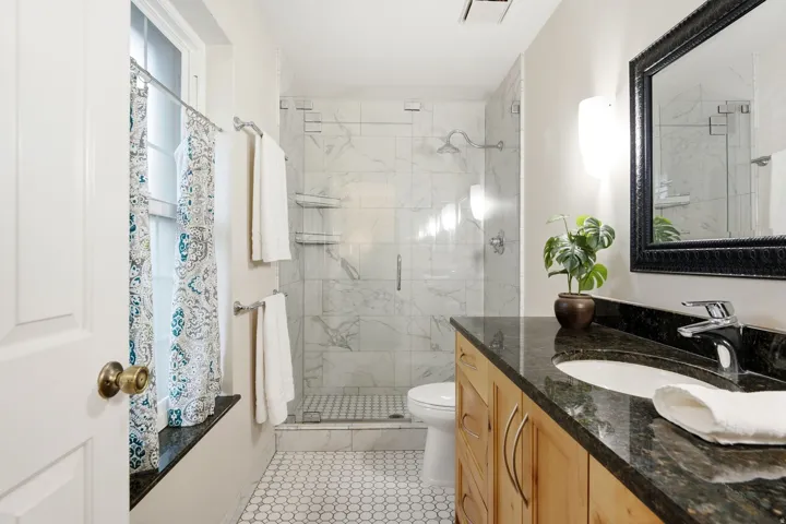 Glass-enclosed shower with white tiled walls, hexagonal floor tiling, natural wood-finish vanity, and dark stone countertop with undermount sink