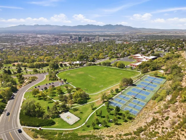 Expansive outdoor recreation area with multiple tennis courts, a large sports field, and paved pathways, complemented by a distant city skyline and mountain backdrop