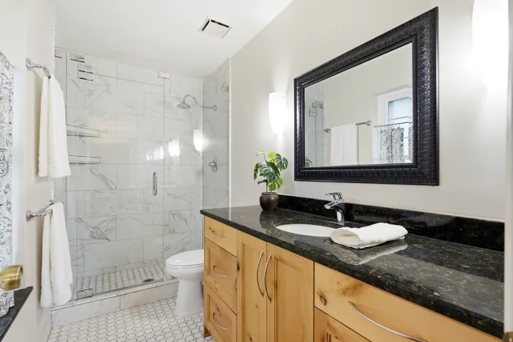 Glass-enclosed shower with white marble-style tile, hexagonal mosaic floor tile, vanity with wood-finish cabinetry, dark stone countertop, and an inset sink