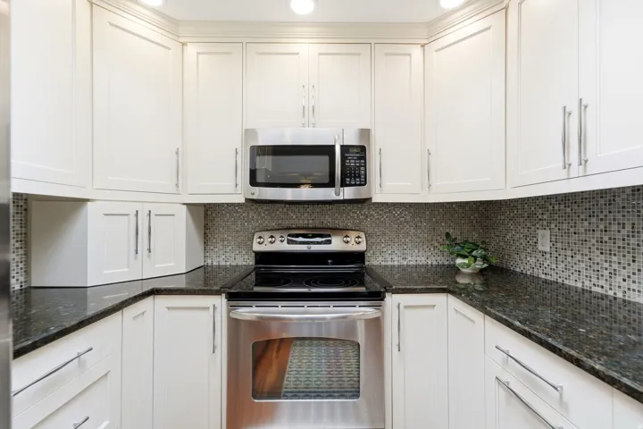 White cabinetry with brushed nickel hardware, dark stone countertops, mosaic tile backsplash, stainless steel microwave, and stainless steel range