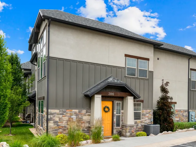 Multi-level residence featuring a stone and stucco facade with vertical paneling, an asphalt shingle roof, and a covered entry with an orange door