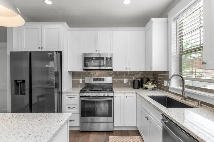 Kitchen featuring white shaker-style cabinetry, light-toned granite countertops, a dark subway tile backsplash, stainless steel appliances, and wood-finish flooring