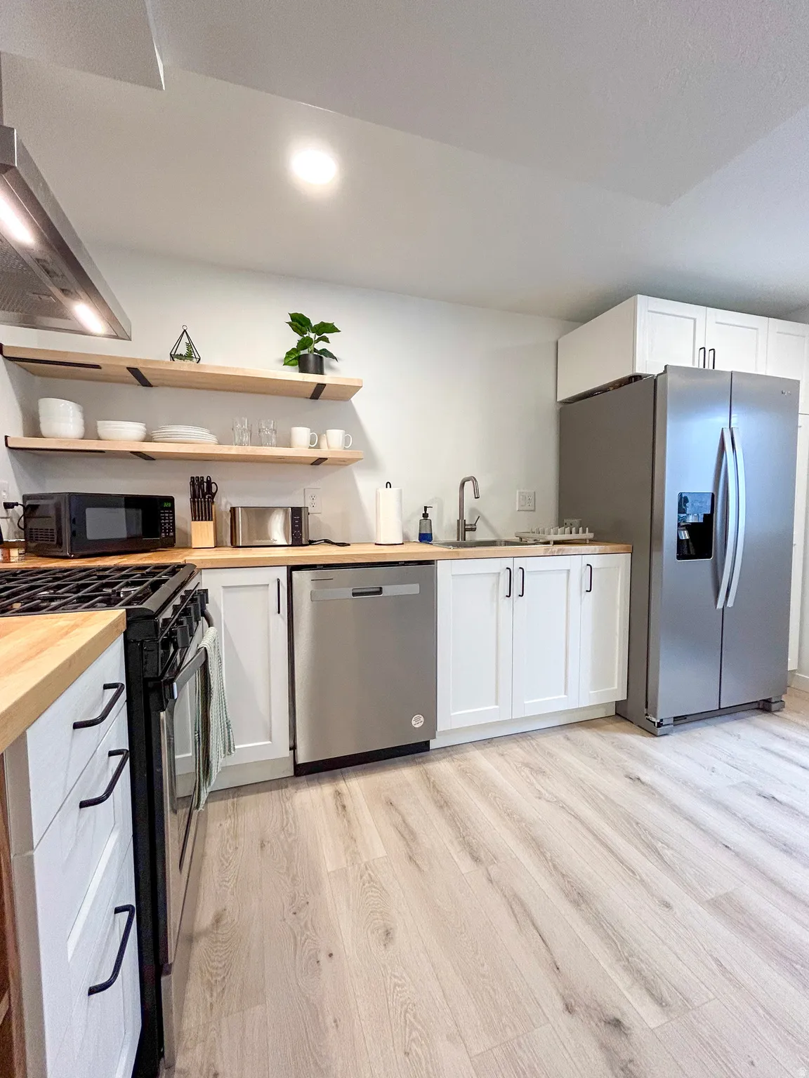 Contemporary kitchen featuring light wood-finish flooring, white cabinetry with black hardware, butcher block countertops, stainless steel appliances, and open shelving