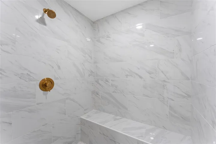 Shower enclosure featuring large format white tiled walls with grey veining, a built-in tiled bench, a bronze finish shower head, and a matching bronze finish shower control