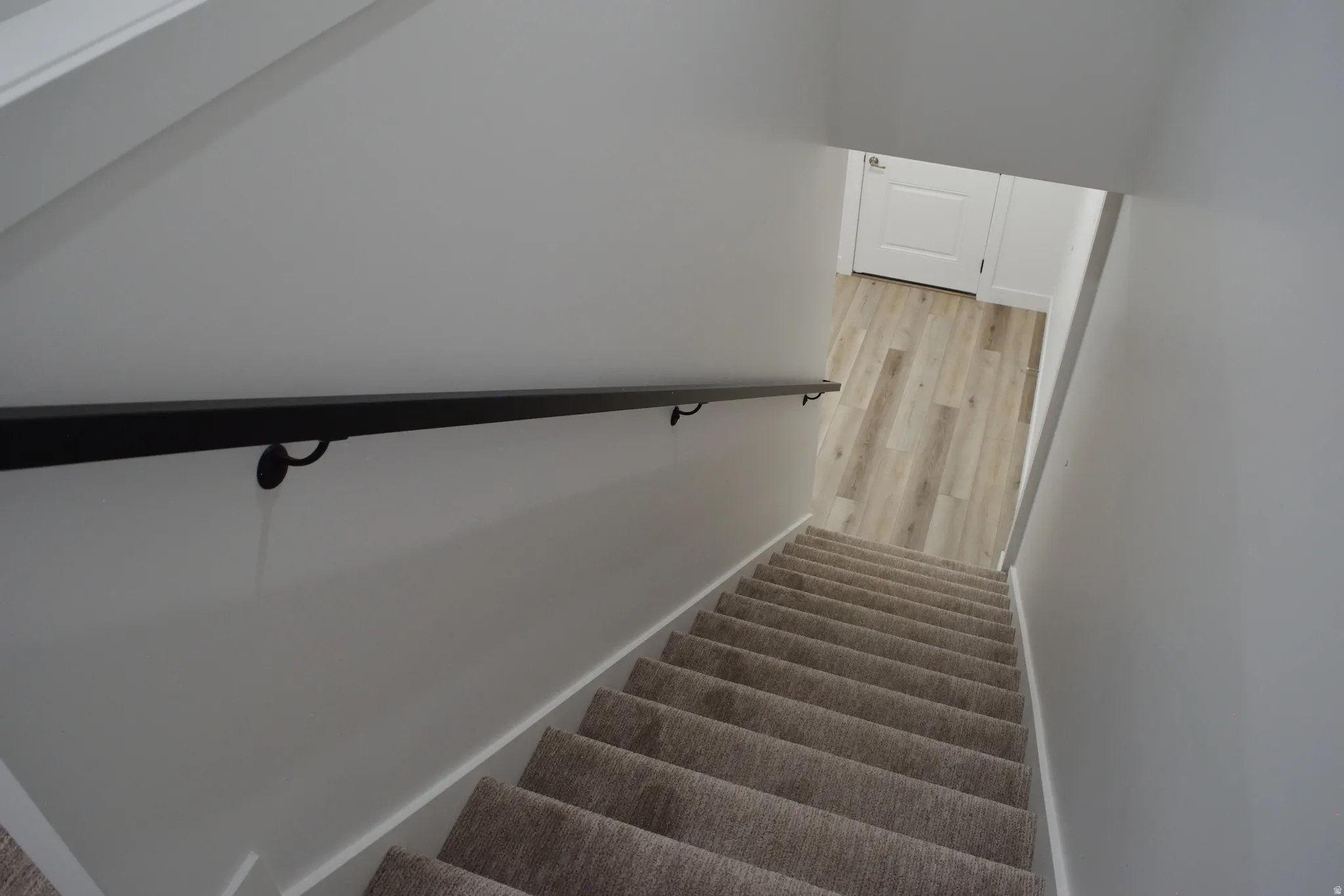 Carpeted staircase with a matte black handrail, leading to a lower level featuring wood-finish flooring and a white paneled door