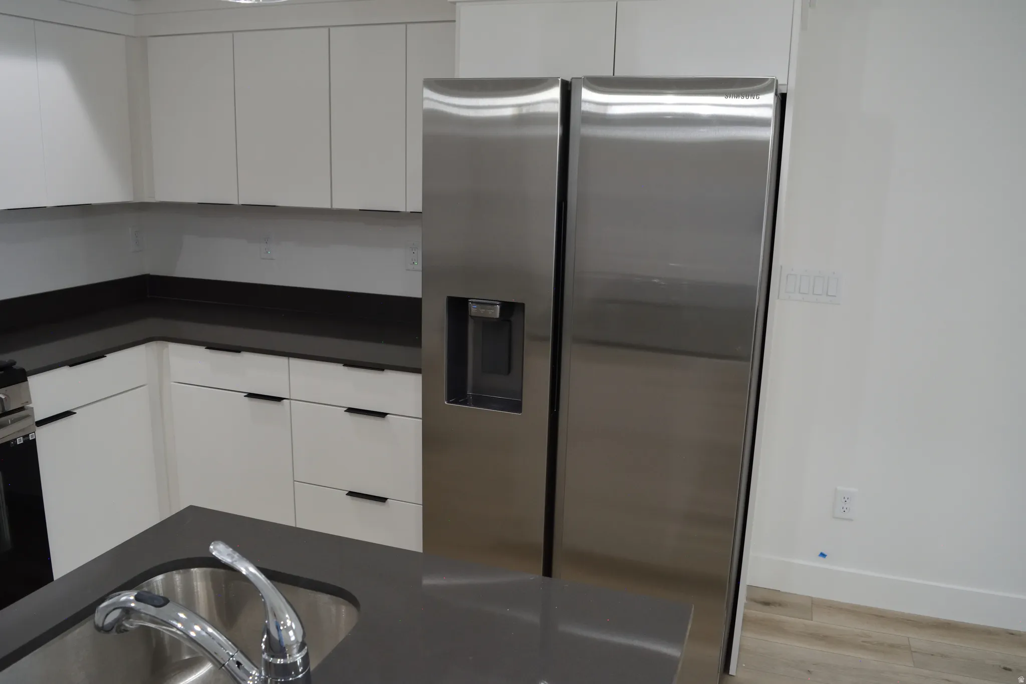 Kitchen featuring white cabinetry with dark pulls, dark countertops, a stainless steel side-by-side refrigerator, a stainless steel sink with a chrome faucet, and wood-finish flooring