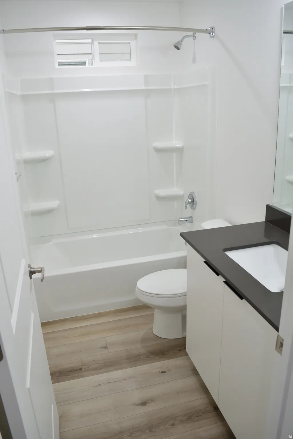 Bathroom featuring wood-finish flooring, a white vanity with dark countertop, an integrated sink, a white toilet, and a bathtub with shower kit