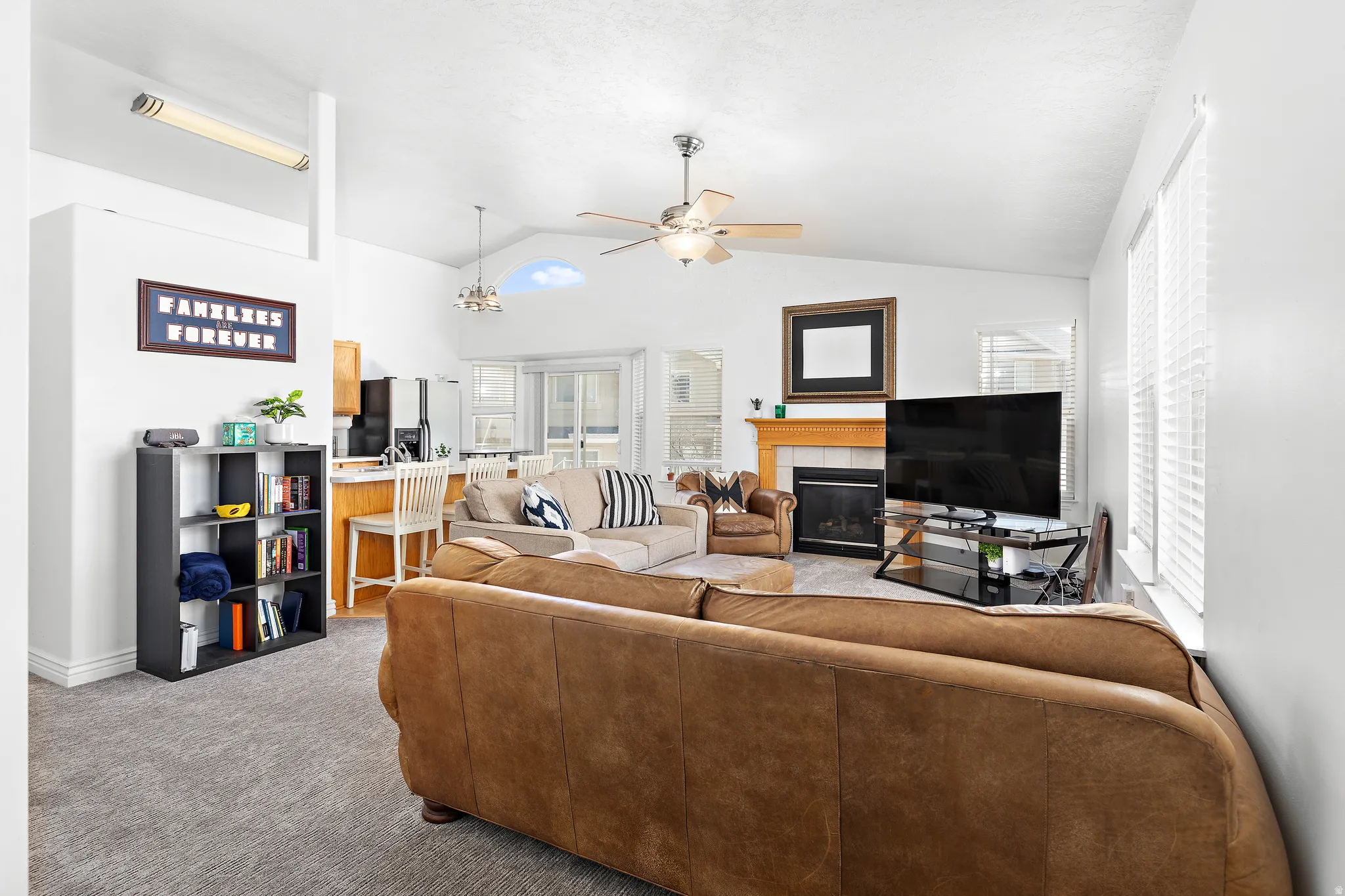 Open-concept living space featuring vaulted ceilings, a fireplace with a wood mantle, a ceiling fan, and large windows with blinds