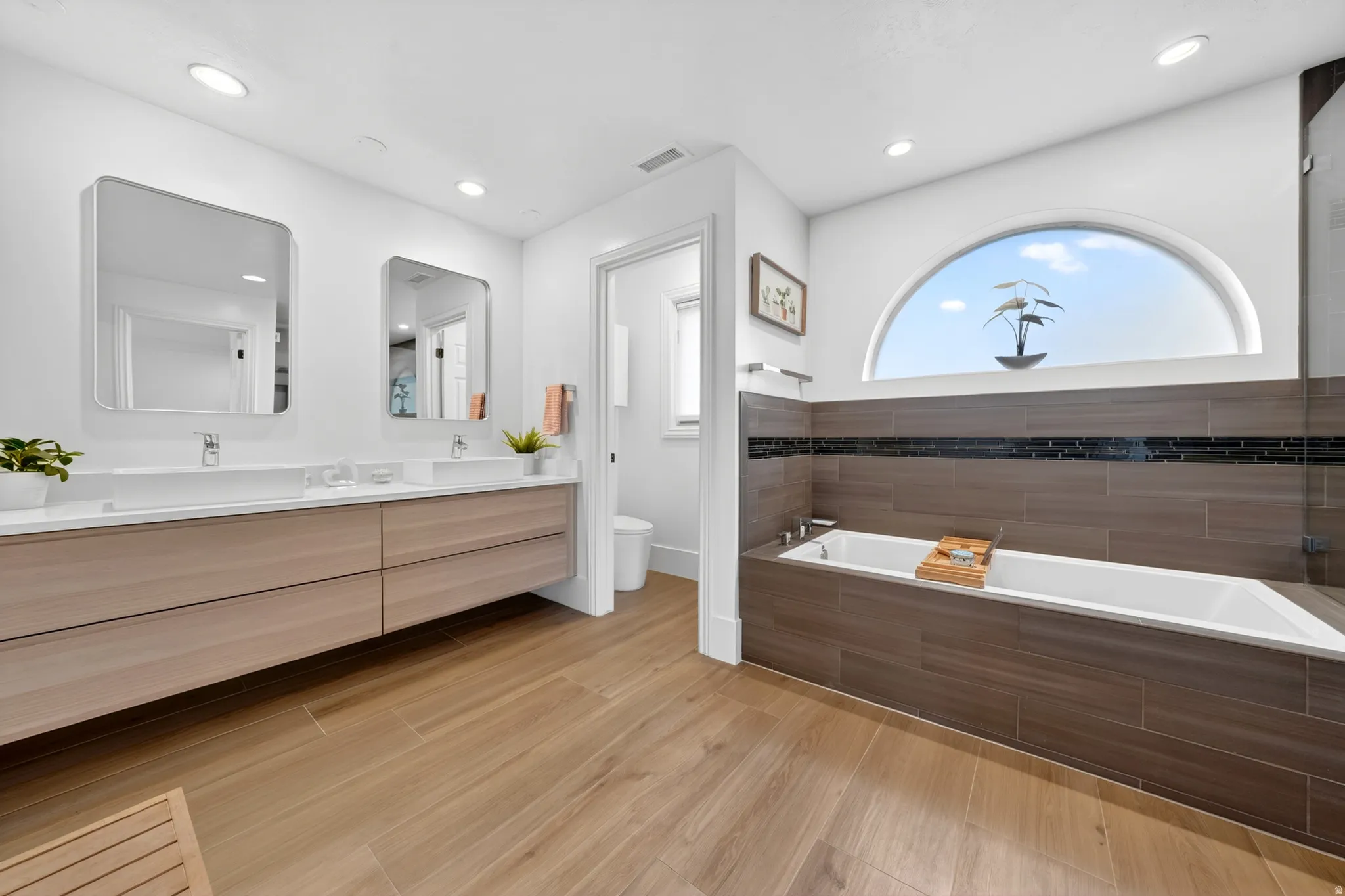 Modern bathroom featuring a dual vanity with light wood-finish cabinetry, a freestanding bathtub, and a large arched window