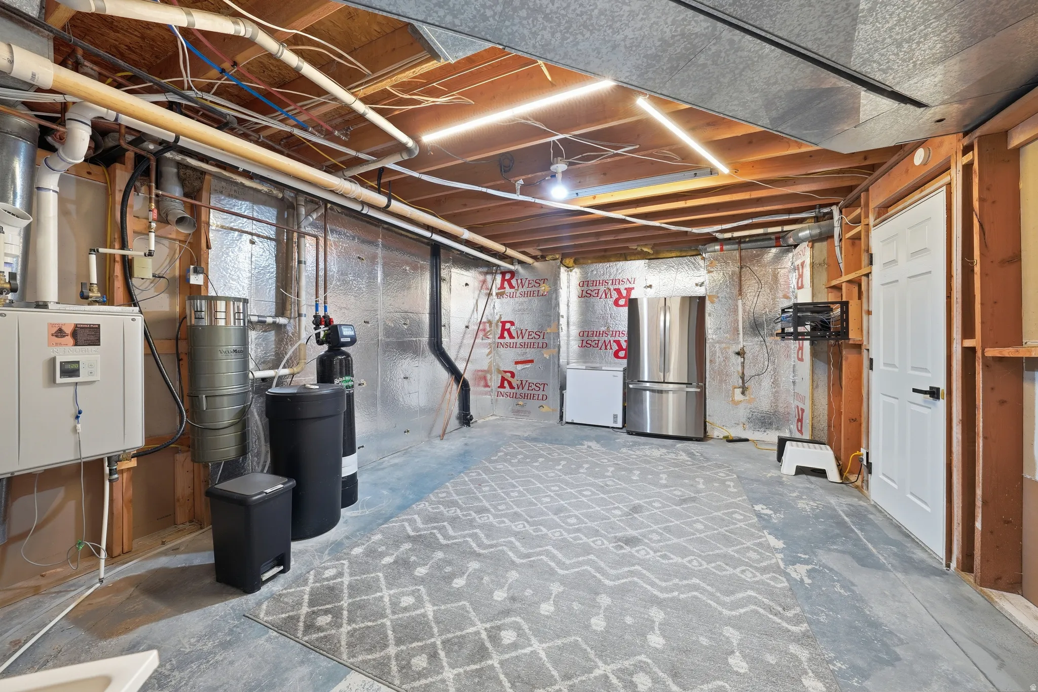 Unfinished basement featuring exposed wood joists, concrete flooring, and visible utility infrastructure including plumbing and electrical conduits