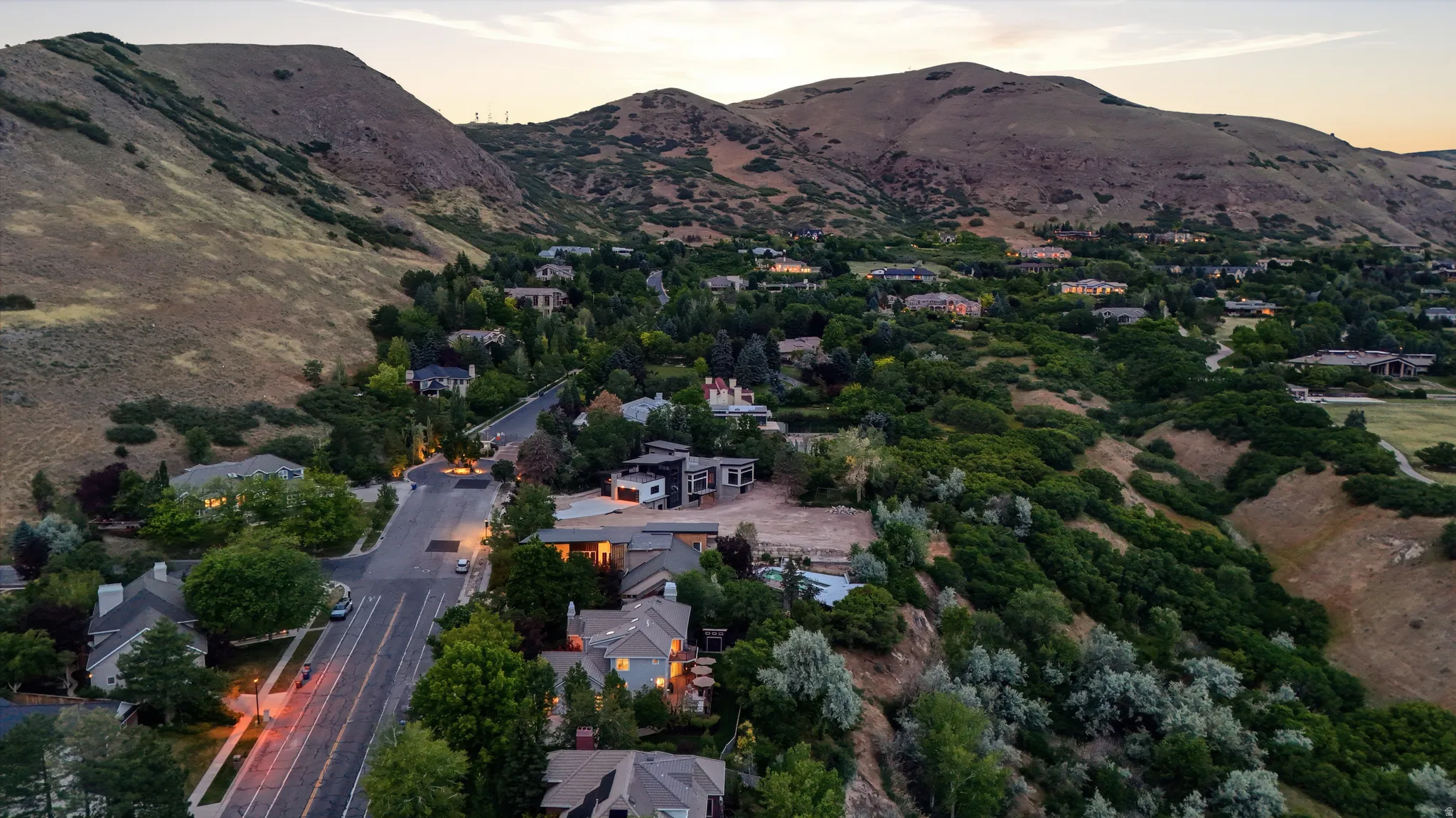 Residential neighborhood nestled against a mountainous backdrop, featuring mature trees and streetlights illuminating the roadway
