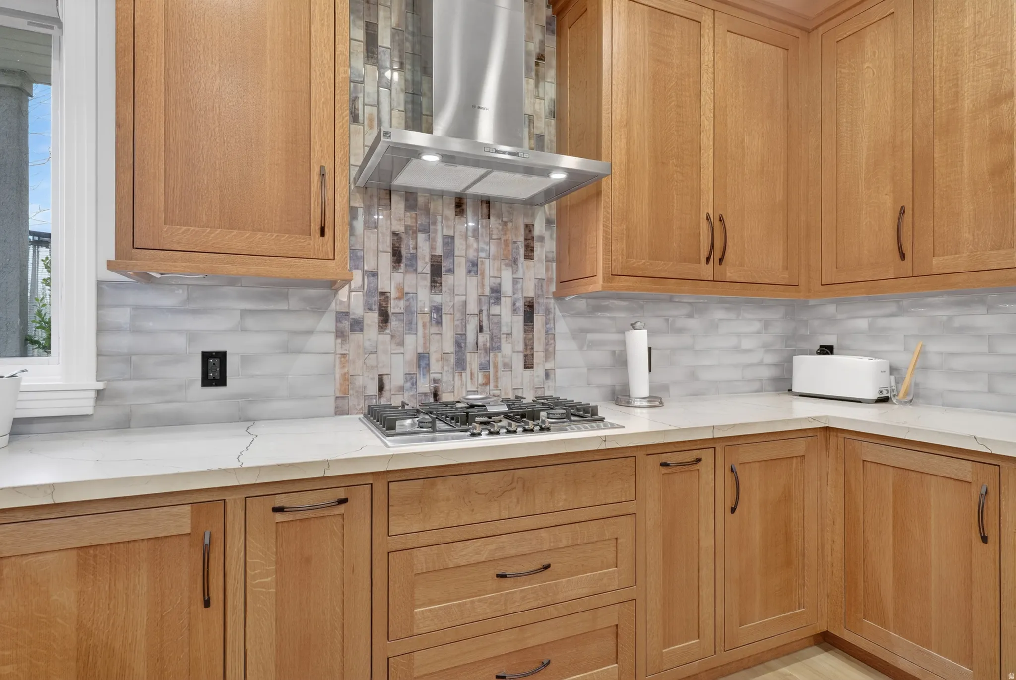 Kitchen featuring light wood cabinetry, white stone countertops, a stainless steel range hood, a gas cooktop, and a mosaic tile backsplash