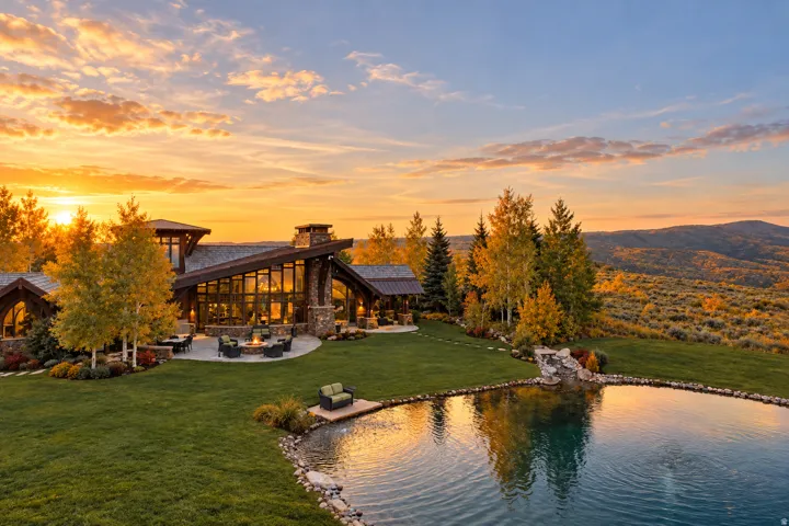 Expansive property featuring a large pond with a waterfall, a stone patio with a fire pit, extensive landscaping, and a striking architectural design with full-height windows