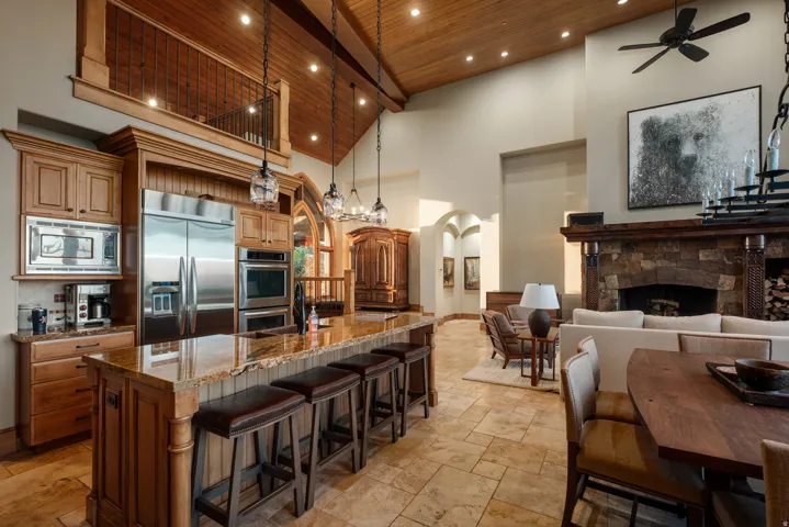 Spacious great room featuring a chef's kitchen with stainless steel appliances, a large center island with seating, a stone fireplace, and a vaulted wood-plank ceiling with exposed beams