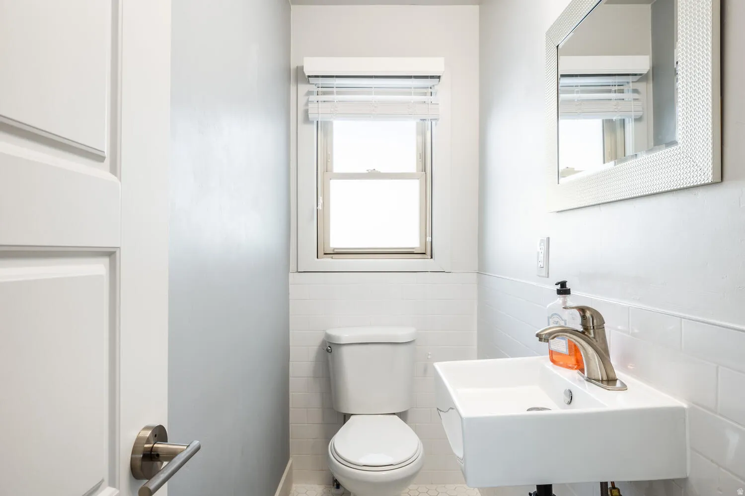 Powder room featuring a wall-mounted sink with brushed nickel faucet, a toilet, a window with horizontal blinds, and white subway tile wainscoting
