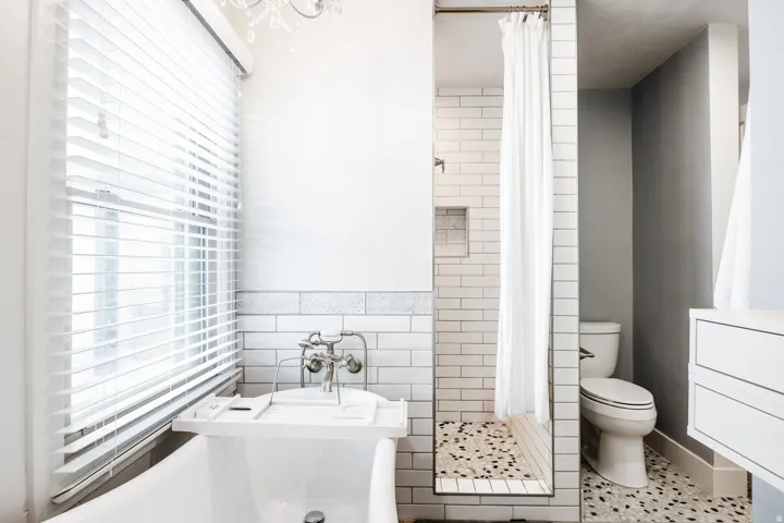 Clawfoot tub with wall-mounted faucet, subway tile wainscoting, and a white wood-slat window blind