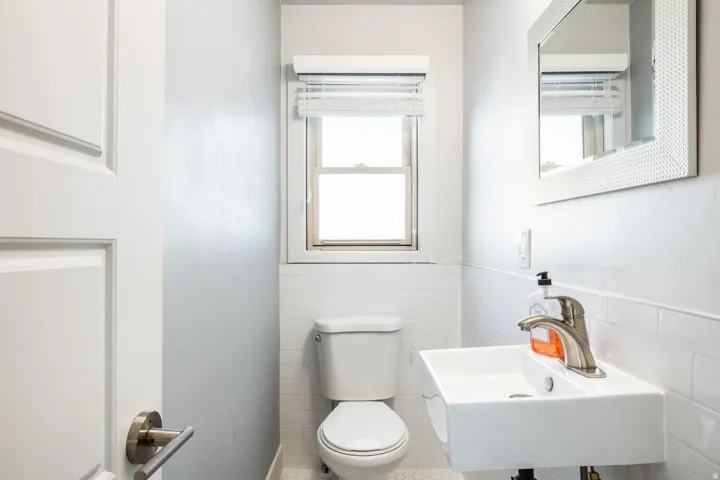 Powder room featuring a wall-mounted sink with brushed nickel faucet, a toilet, a window with horizontal blinds, and white subway tile wainscoting