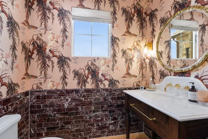 Powder room featuring botanical-patterned wallpaper, dark marble subway tile wainscoting, and a white vanity with gold-toned fixtures