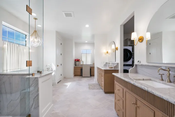 Spacious bathroom featuring light-toned flooring, dual vanity areas with stone countertops and light wood cabinetry, a walk-in shower with clear glass enclosure and stone surround, and a dedicated laundry area with stacked appliances