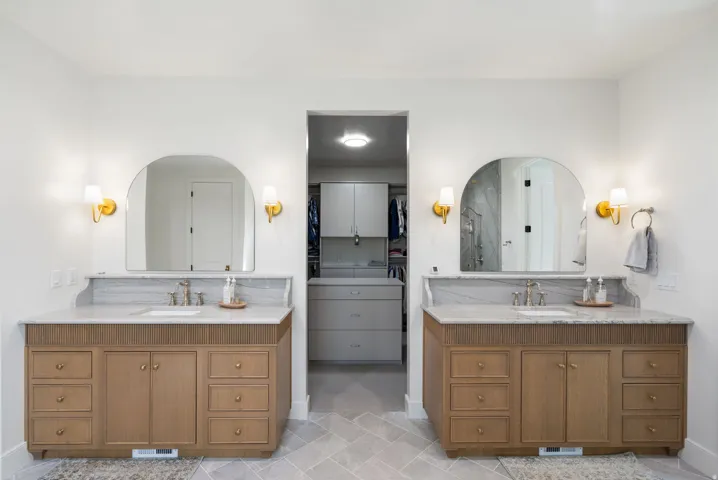 Dual vanity with light wood-finish cabinetry, integrated sinks, and polished countertops, each featuring arched mirrors and brass sconces