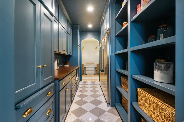 Expansive pantry featuring custom blue cabinetry, open shelving, brass hardware, a wood-finish countertop, and checkered tile flooring