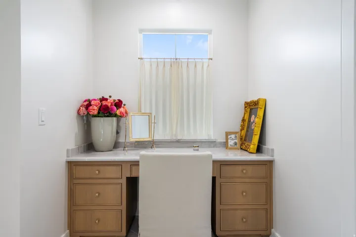 Built-in vanity with a light wood-finish and brass hardware, featuring a stone countertop and a window with sheer curtains