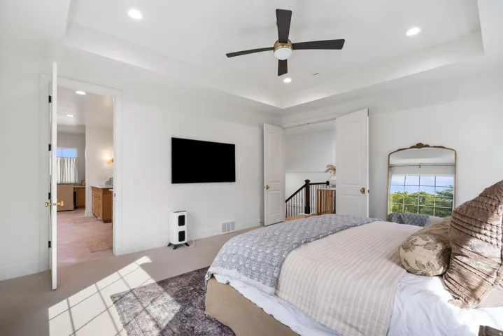 Spacious bedroom featuring a tray ceiling with recessed lighting and a ceiling fan, a wall-mounted television, and neutral carpeting