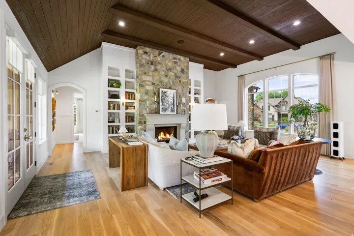 Spacious living area featuring a stone fireplace with a substantial mantel, built-in shelving, wood-finish flooring, a wood-paneled vaulted ceiling with exposed beams, and large arched windows